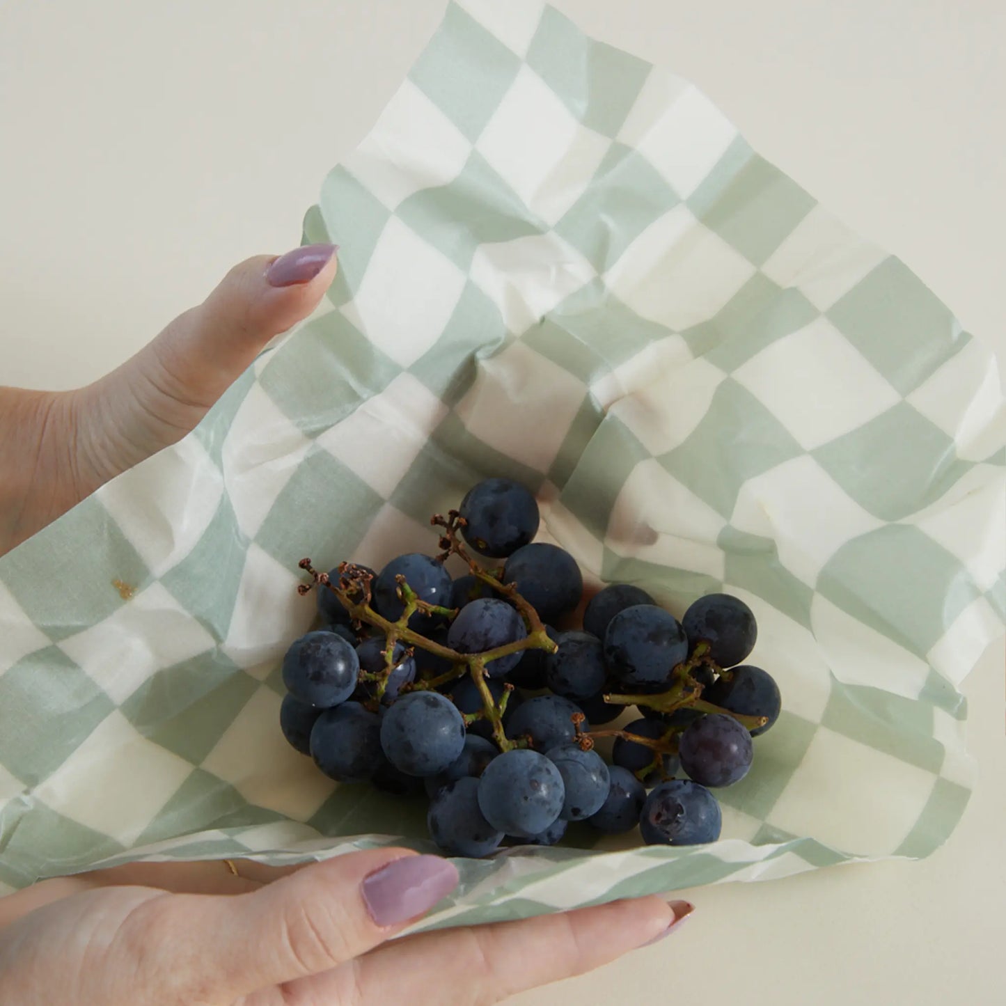 A person holds a bunch of dark grapes wrapped in an eco-friendly beeswax wrap from the Beeswax Wrap Set of 3 - Geo, featuring a light green and white checkered pattern.