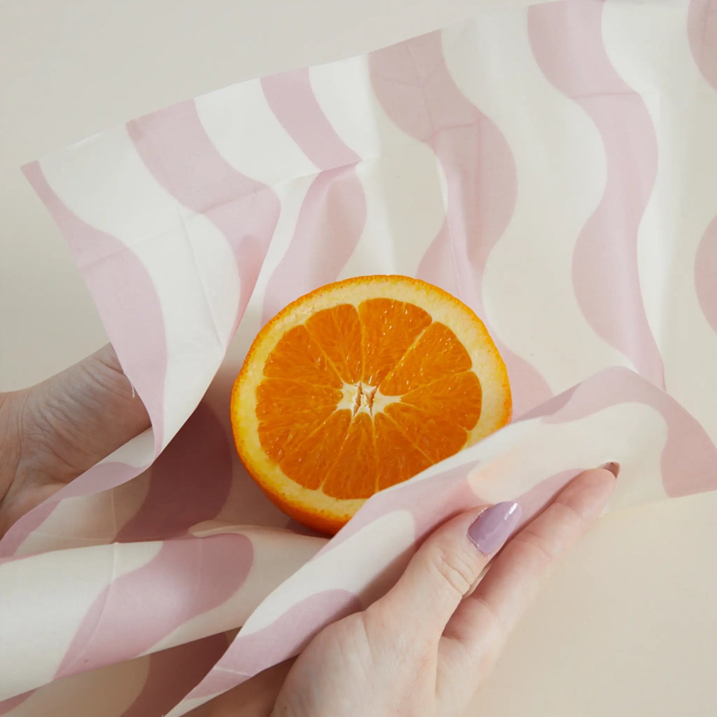 A person with painted nails wraps half an orange in a pink and white Beeswax Wrap Set of 3 - Geo, highlighting an eco-friendly alternative to plastic.