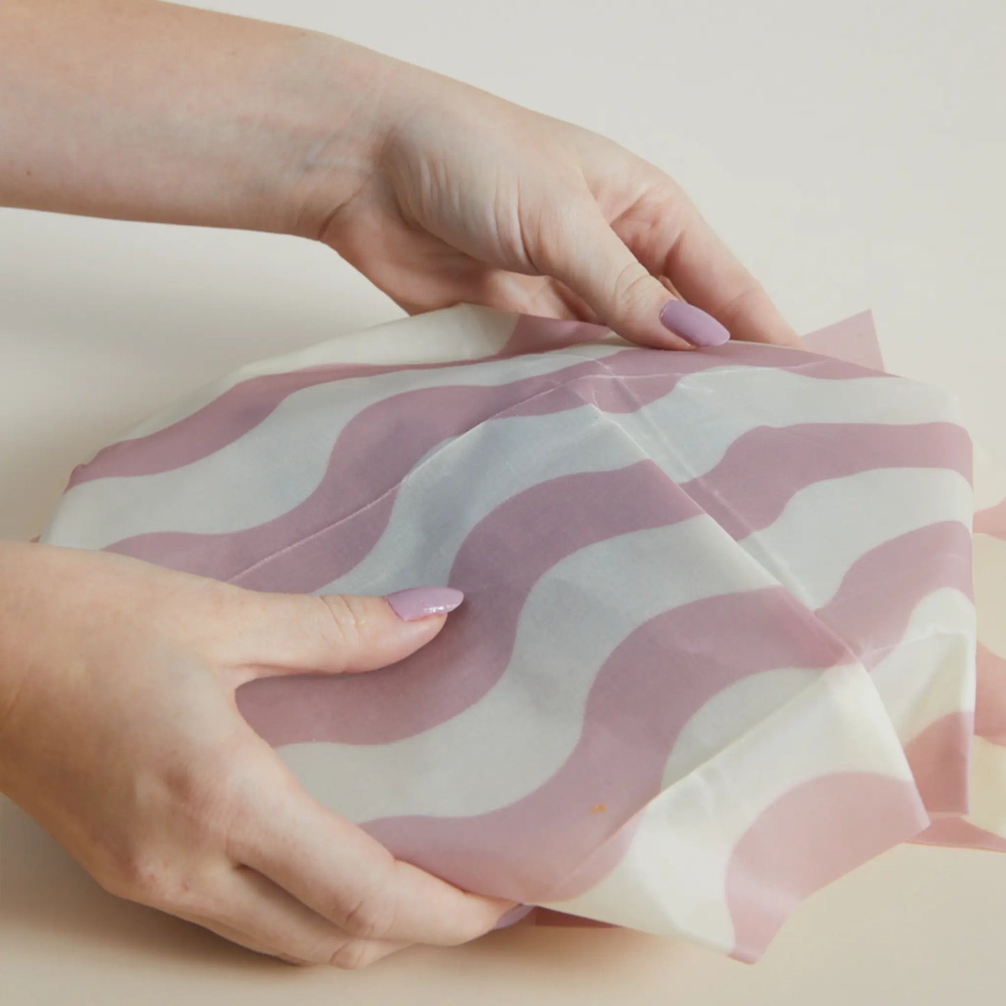 Two hands place a Beeswax Wrap Set of 3 - Geo, featuring pink and white wavy stripes, over a bowl. This eco-friendly, reusable food wrap helps reduce waste and keeps food fresh.