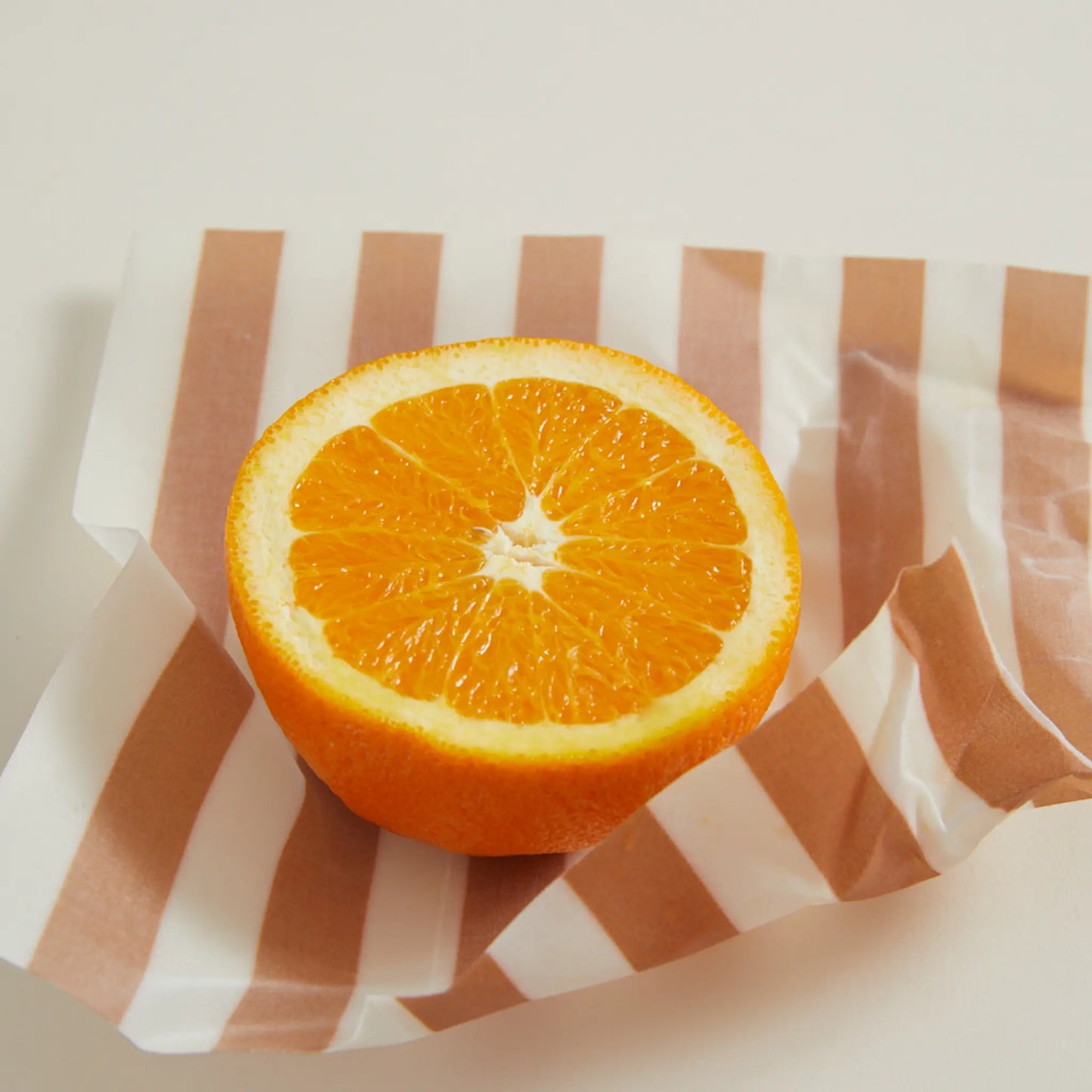 A halved orange sits on a pink and white striped Beeswax Wrap Set of 3 - Geo, an eco-friendly alternative to plastic, atop a light-colored surface.