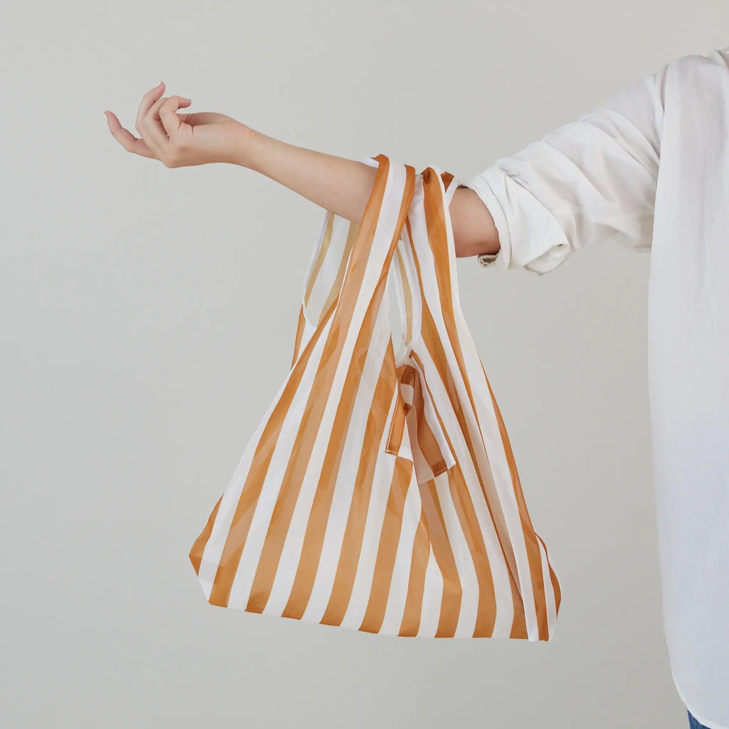 A person wearing a white shirt holds the Reusable Market Bag - Stripe, featuring orange and white stripes, on their arm against a plain background.