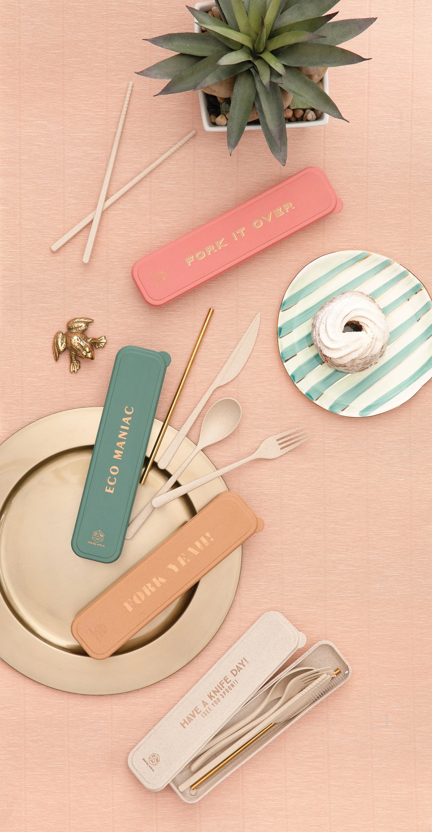 A flat lay of the Have A Knife Day! (See You Spoon!) Portable Flatware Set in vibrant eco storage cases, gold utensils on a plate, striped dish with pastry, chopsticks, and a small potted succulent on a pink surface.