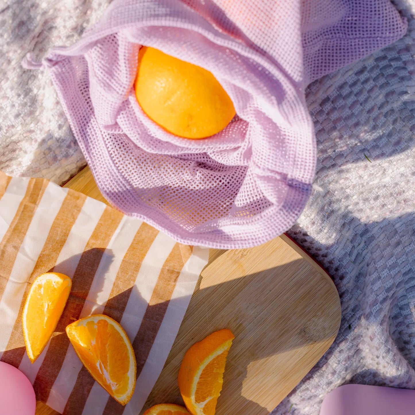 A lilac Reusable Produce Bag from the Rust, Lilac, & Hunter set holds an orange on a textured blanket beside a wooden cutting board with sliced oranges, highlighting stylish and eco-friendly produce storage.