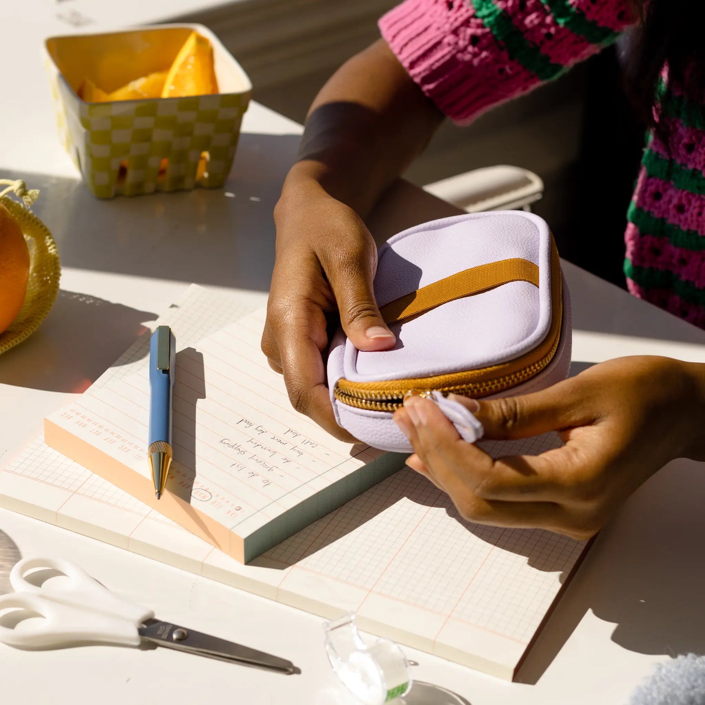 Someone holds the Office On The Go - Lilac + Copper vegan leather zippered pouch above a notebook, surrounded by a pen, scissors, washi tape, and orange slices—ideal for staying organized at work.