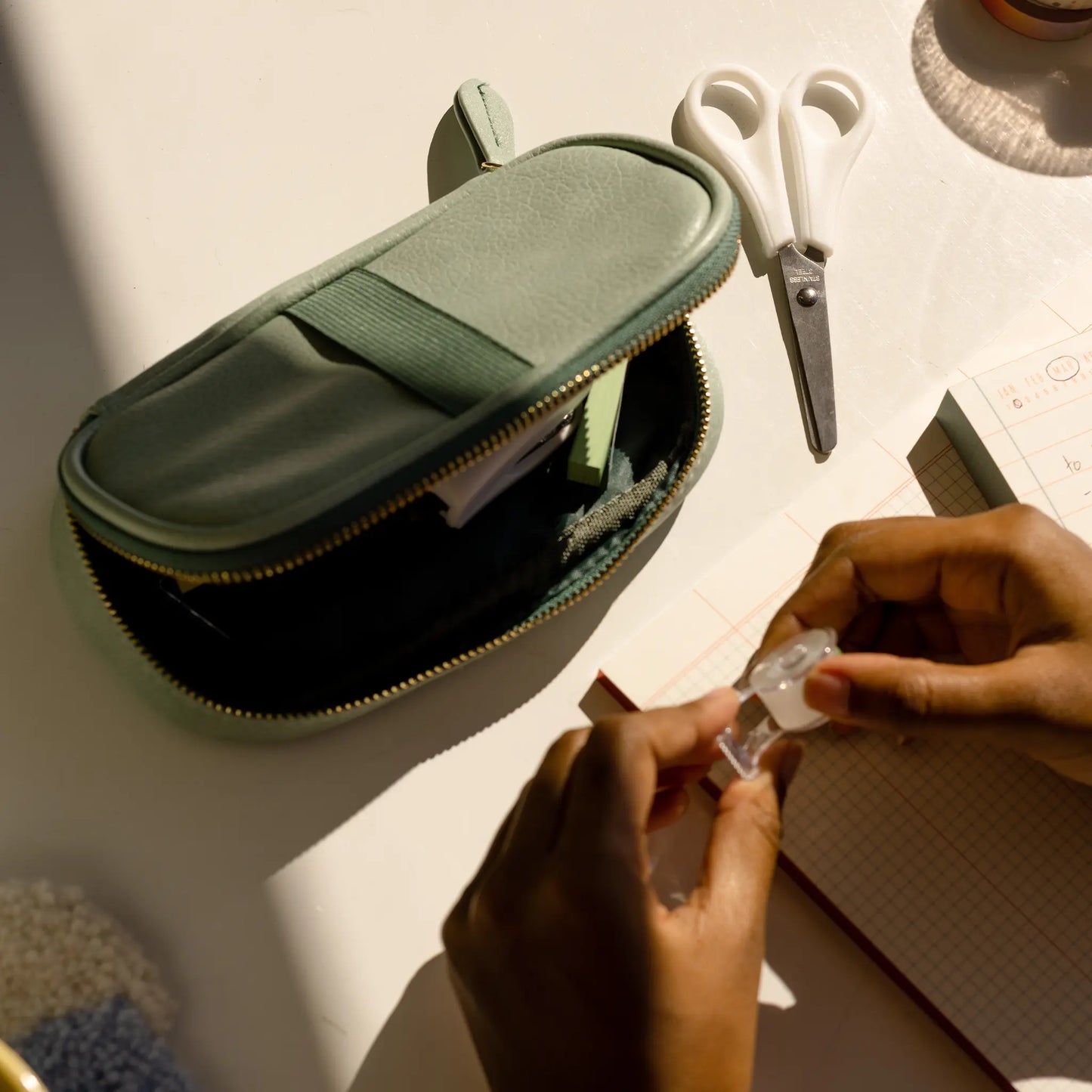 A person holds correction tape at a desk with an open Office On The Go - Matcha + Forest vegan leather pencil case, white scissors, and a notebook.
