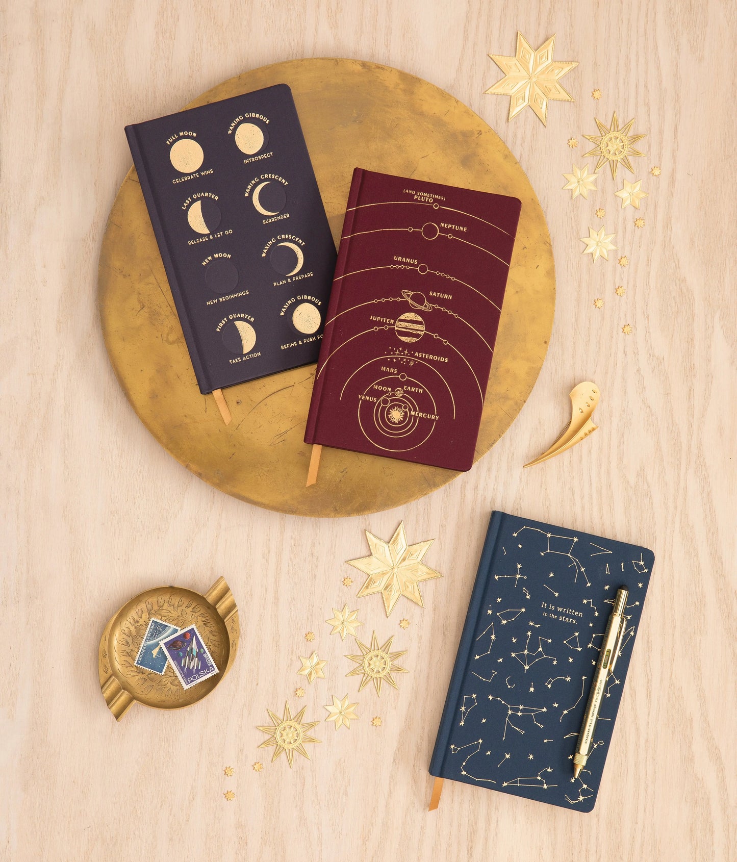 The Cloth Journal - Solar System collection includes two burgundy journals—one with moon phases, one with planetary orbits—and a navy journal with constellations. All are arranged on wood with gold accents and feature acid-free paper for durability.