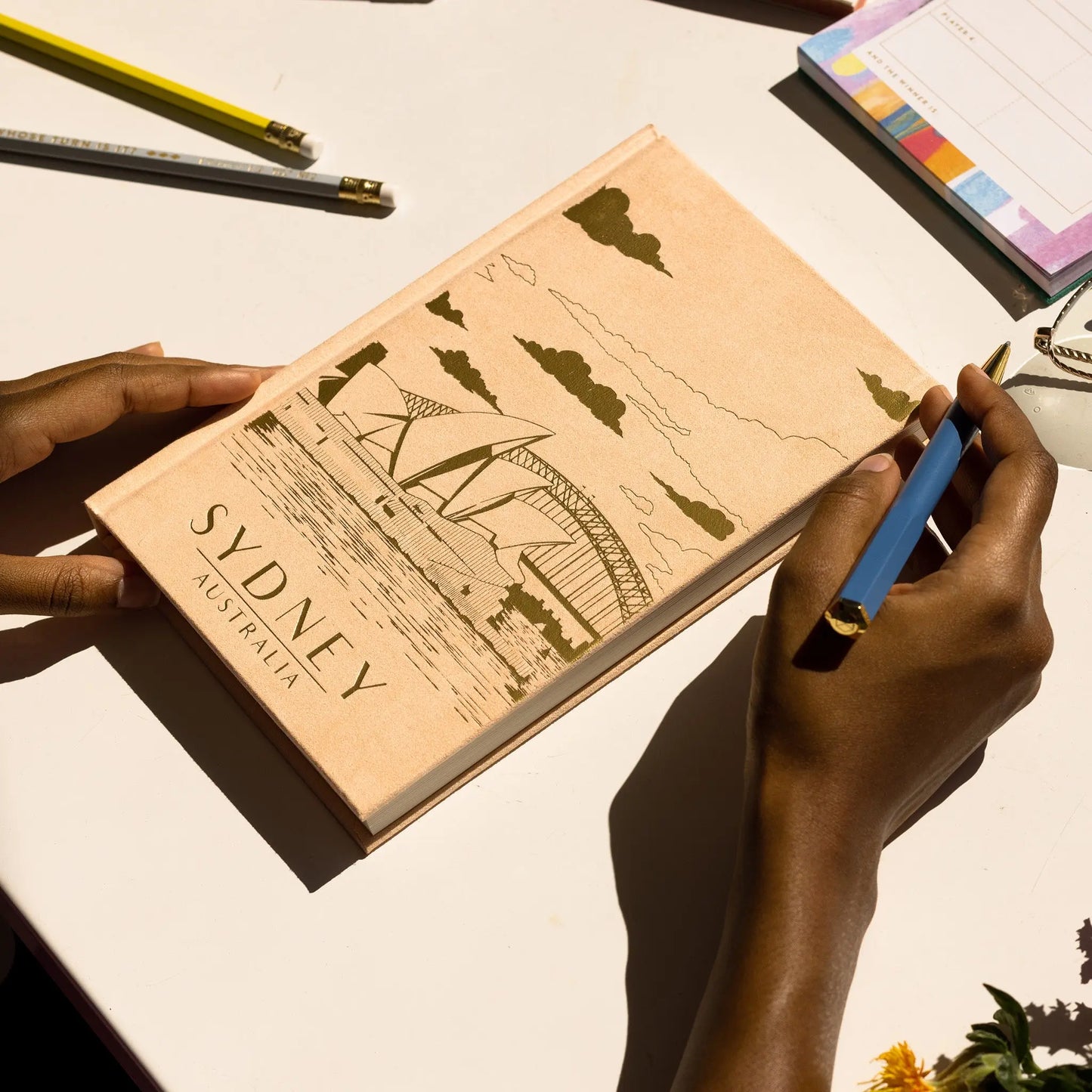 Person holding an Anderson Design Journal - Sydney, a beige sueded paper journal with a vintage poster of the Sydney Opera House and Harbour Bridge; pencils, a notepad, and flowers are arranged on the white table.