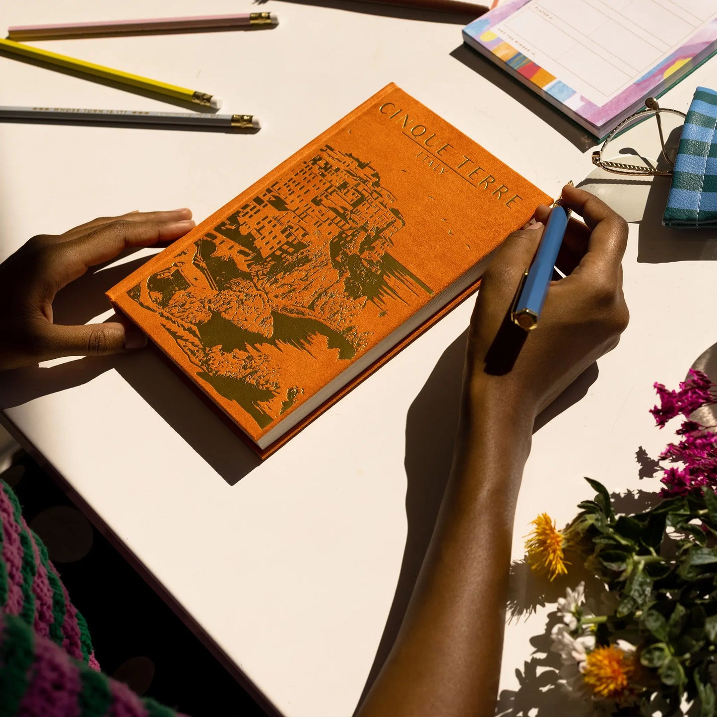 A person holds the Anderson Design Journal - Cinque Terre, featuring subtle vintage poster design, at a desk with stationery, flowers, and a notepad—capturing the charm of the Italian Riviera.