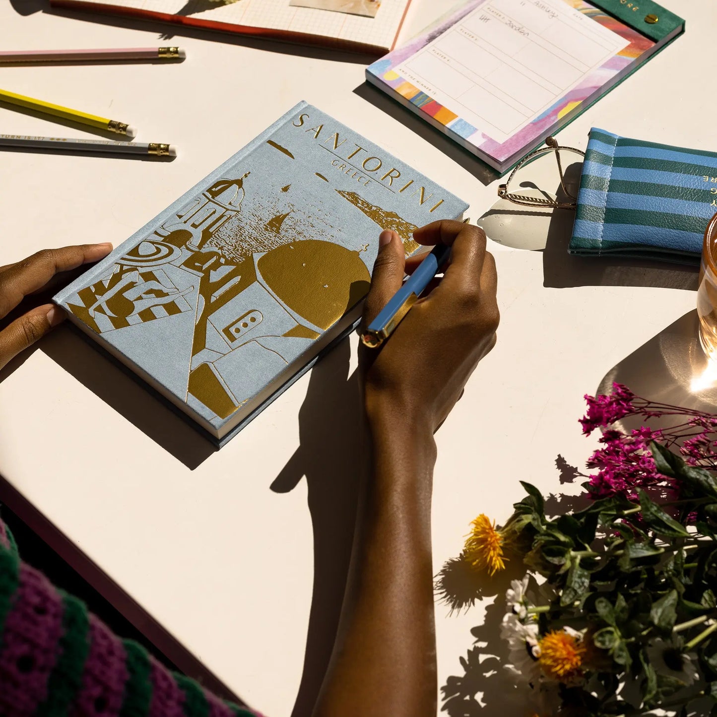 At a desk with flowers, pens, a notepad, and a striped pouch, someone writes in the Anderson Design Journal - Santorini, featuring sueded blue paper and capturing the nostalgic charm of vintage poster art.