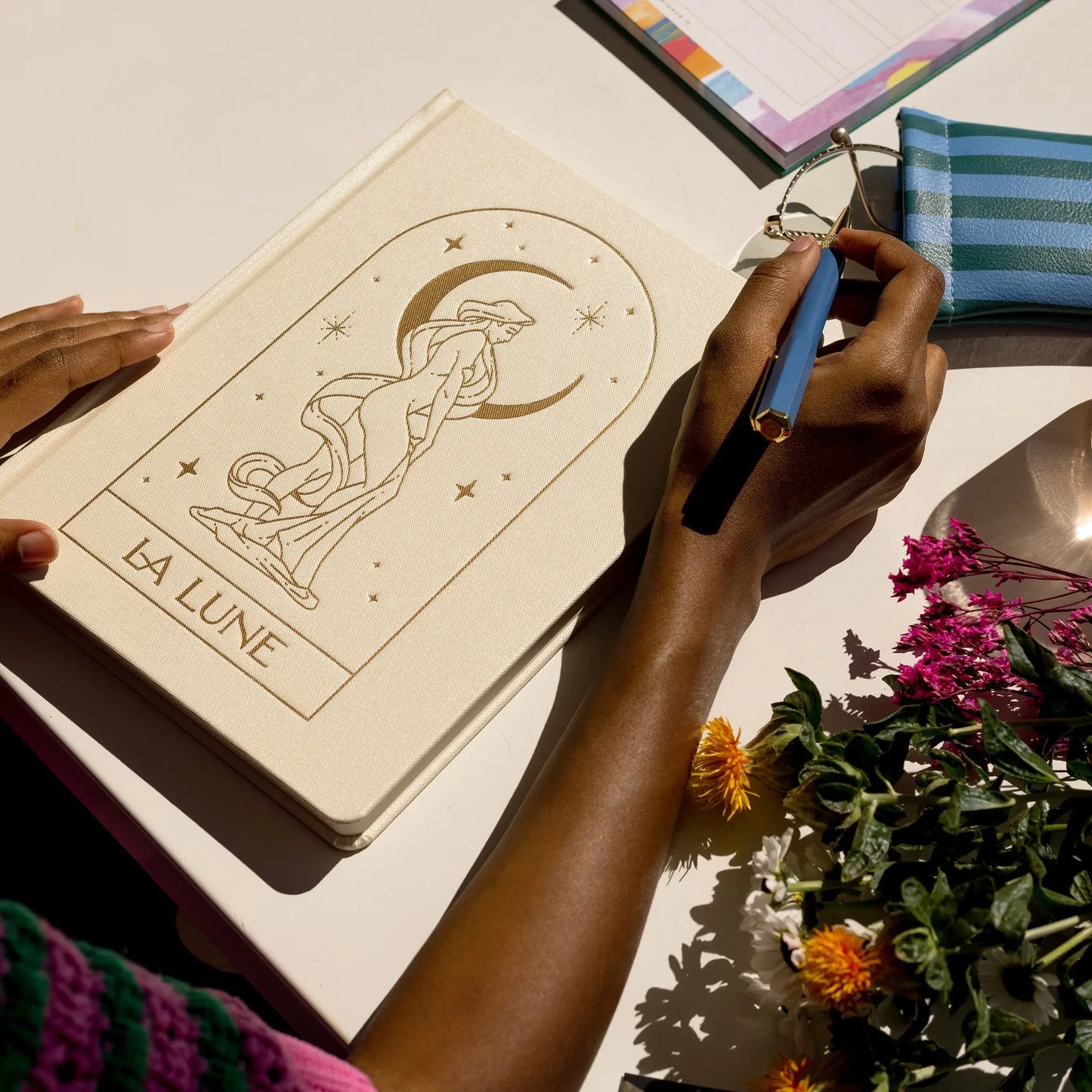 A person writes with a pen beside the Bookcloth Jumbo Journal - La Lune, assorted flowers, a striped pouch, and a notepad on a white desk, enjoying the smooth touch of acid-free paper.