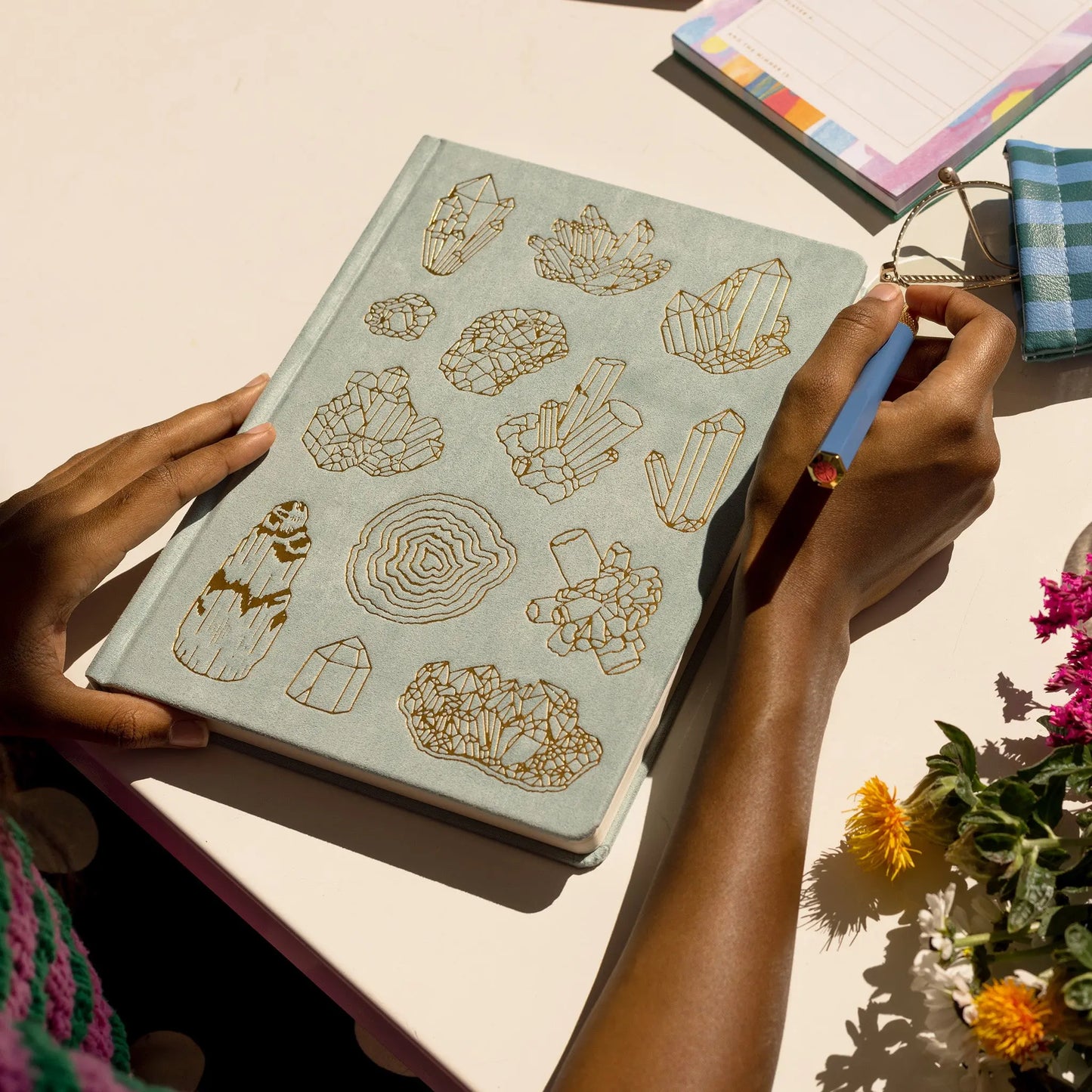 A person holds a pencil near the Suede Jumbo Journal - Mineralogy, featuring gold crystal illustrations; flowers, a notepad, and a striped pouch surround the lay-flat binding journal on the table.