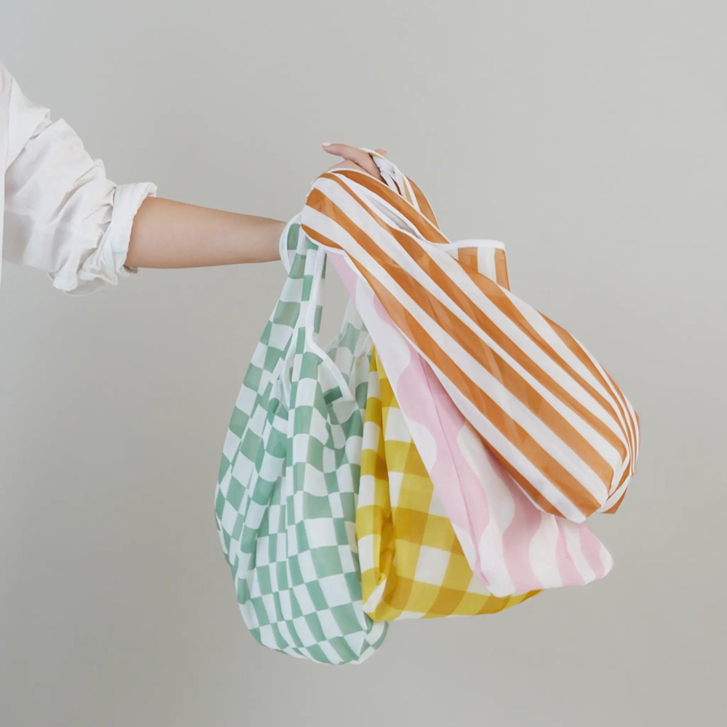 A person holds three Reusable Market Bag - Checkered styles—green checkered, yellow checkered, and orange striped—against a plain background. Made from recycled bottles, these eco-friendly bags are perfect for shopping.