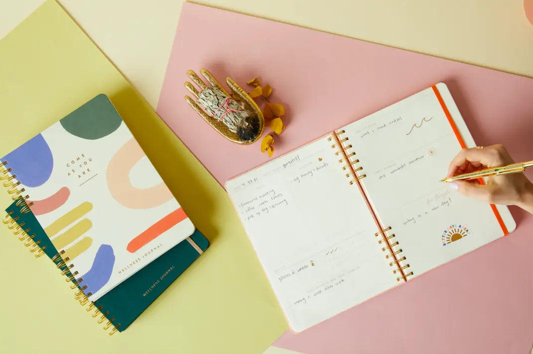 A hand writes in the Take Care Guided Wellness Journal on a desk with a closed notebook, a decorative hand-shaped dish, and colorful papers—ideal tools for your gratitude practice and self-care journey.