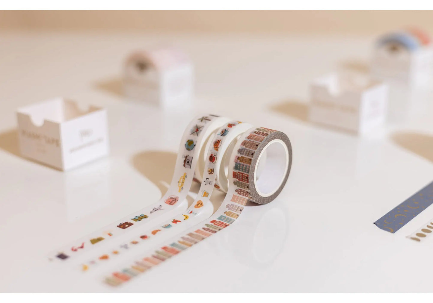 A roll from the Washi Tape Set of 3 - Travel, featuring colorful travel designs, is unrolled on a white surface—ideal for scrapbooking or bullet journals. Blurred tape boxes and other rolls are visible in the background.