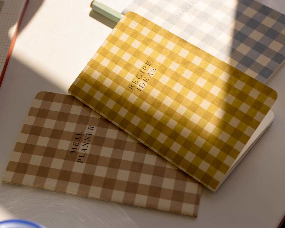Two checkered notebooks labeled MEAL PLANNER and RECIPE IDEAS are placed on a table with sunlight casting shadows across them.