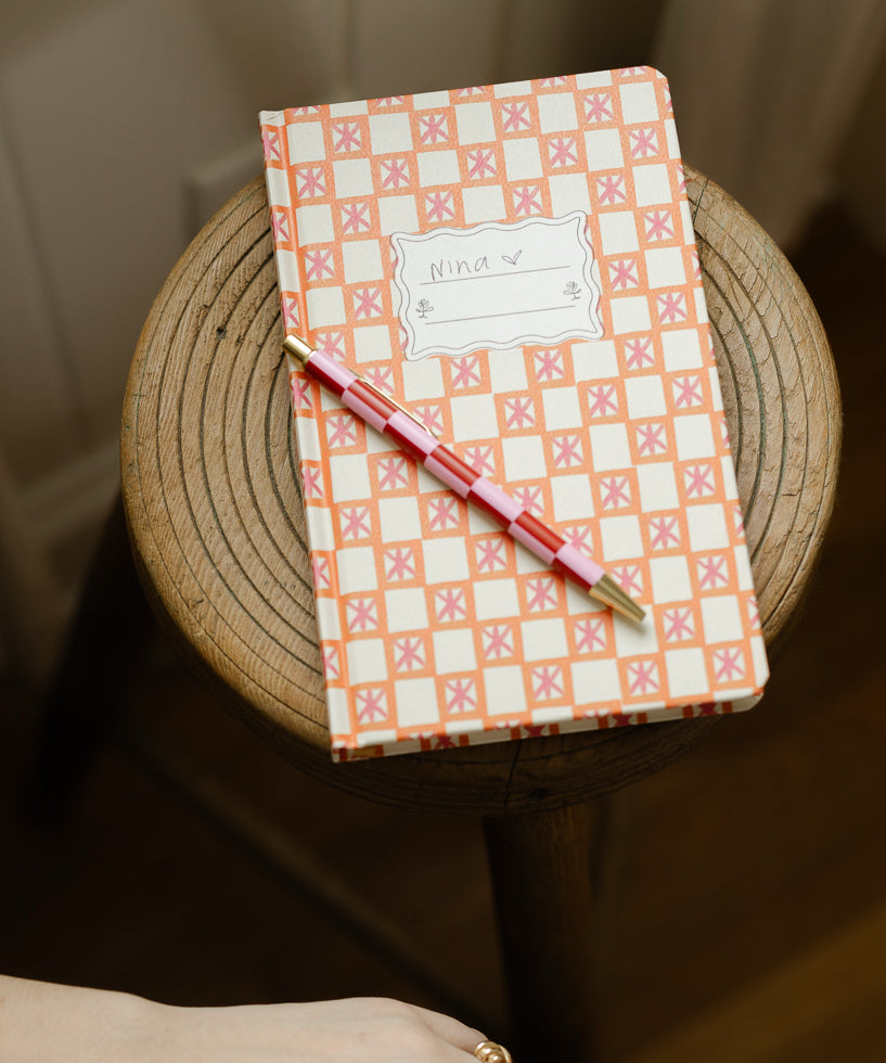 A notebook with a pink and orange checkered cover labeled Nina and a pink pencil rests on top, placed on a round wooden stool.
