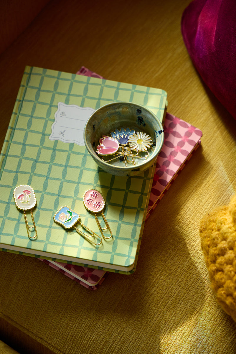 A small bowl of decorative pins sits on two patterned notebooks, with several colorful enamel paperclips lying nearby on the notebooks, in warm lighting.