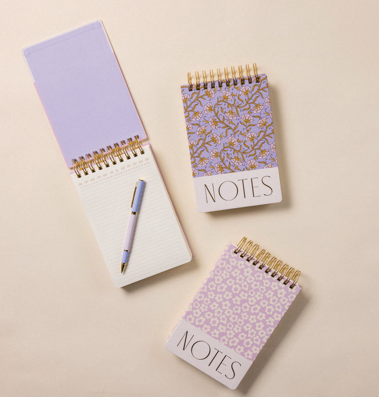 Three spiral-bound notepads with NOTES on the cover, two featuring floral designs, and one open notepad with lined pages and a pen resting on top, all on a cream background.