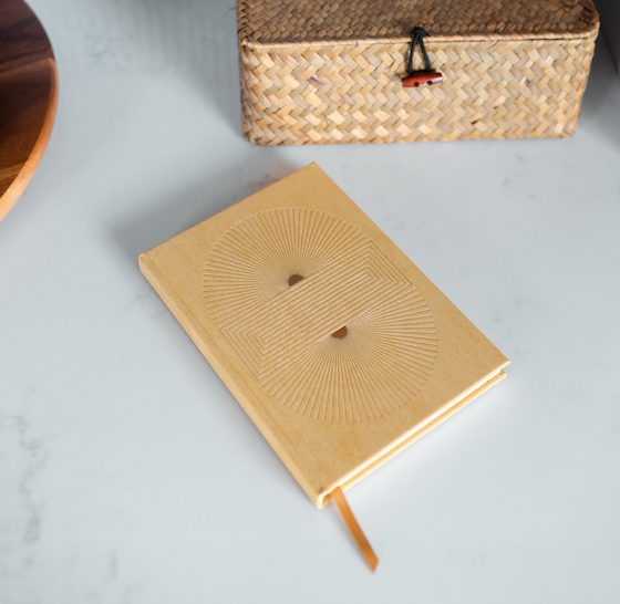 A tan notebook with an embossed geometric design lies closed on a white surface, next to a woven basket and a wooden tray.