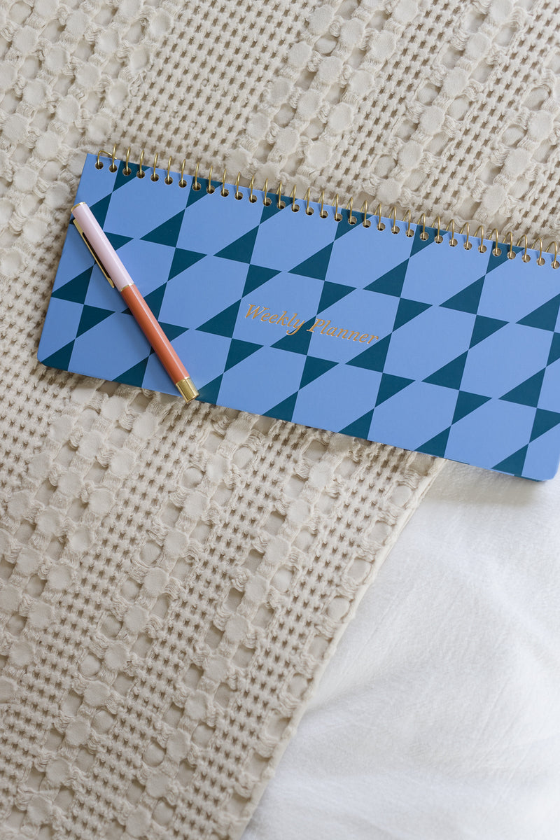 A blue weekly planner with a geometric pattern and gold spiral binding lies on a textured beige blanket, with a pink and white pen resting on top.