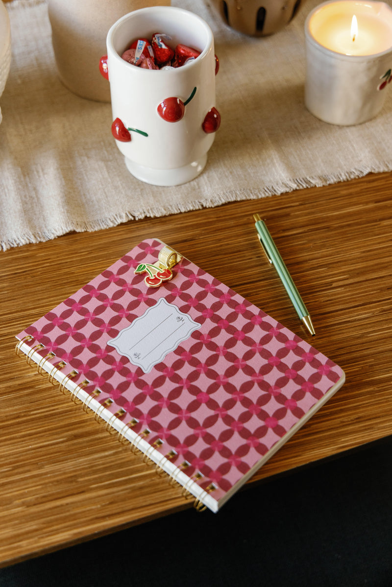 A pink patterned spiral notebook with a blank label sits on a wooden table next to a green pen, a lit candle, and a white cup decorated with cherries and filled with wrapped candies.