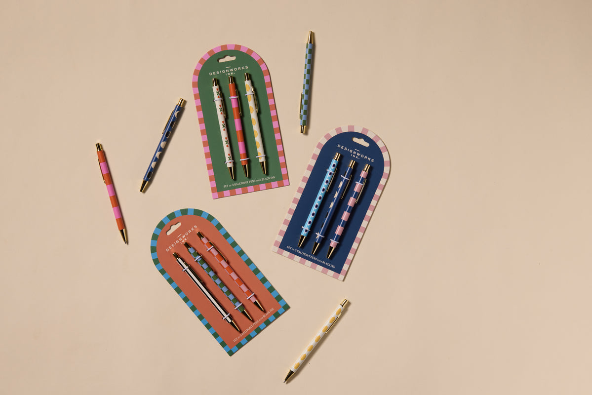 The A Dopo Set Of 3 Pens - Stripes & Checks includes three colorful pens in Mediterranean-inspired packaging, arranged on a beige surface alongside two single pens. Each pen features bright striped and dotted patterns.