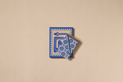 Two blue A Dopo Eye Twin Wire Notepads with hand-drawn geometric and eye patterns are stacked on a beige background, with a larger notebook at the bottom and two smaller spiral notepads on top.