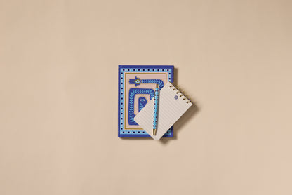 The A Dopo Eye Twin Wire Notepad, with its blue patterned cover and hand-drawn designs, rests on a beige surface alongside a small spiral notepad and blue pencil—ideal for recording Mediterranean travel memories.