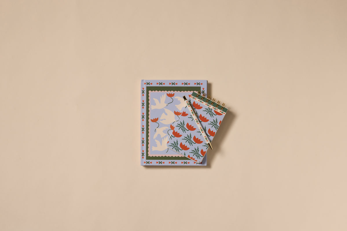 Two A Dopo Flower Journals with decorative covers rest on a beige surface; the larger has a blue and white dove pattern, while the smaller features red birds and green leaves—ideal for journaling or gifting to creatives.