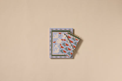 Two creative stationery pieces on beige: a notebook with blue and white doves, and the A Dopo Flower Twin Wire Notepad featuring a blue and red bird among branches. Both are finished with decorative patterned borders.