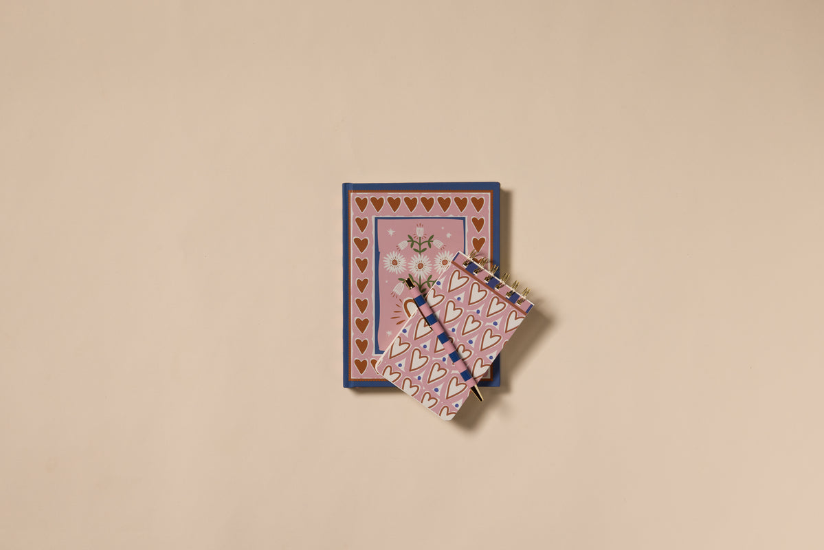 Two A Dopo Hearts Journals are stacked on a beige background. The larger has a soft-touch cover with gold hearts and daisies, while the smaller is decorated with pink and gold hearts for extra charm.