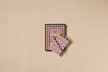Two A Dopo Hearts Journals are stacked on a beige background. The larger has a soft-touch cover with gold hearts and daisies, while the smaller is decorated with pink and gold hearts for extra charm.