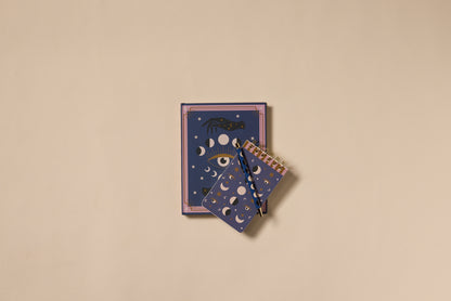 The A Dopo Moon Phases Twin Wire Notepad features gold moon phases, stars, and a hand-drawn eye on dark blue covers. Two notepads rest on a beige background, with one spiral-bound notepad overlapping the larger notebook beneath.