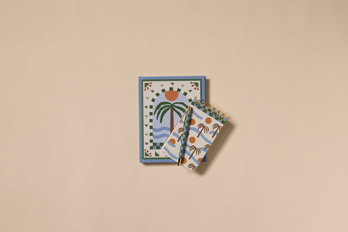 Two lay-flat notebooks with abstract nature designs rest on a beige background. The larger, the A Dopo Palm Journal, features a palm tree and sun; two smaller spiral travel journals display playful palm patterns.
