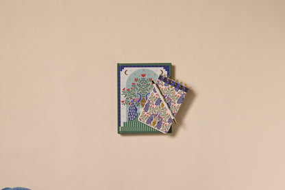 Two A Dopo Peacock Journals with vibrant floral and tree designs, featuring soft-touch covers—one spiral-bound—are stacked on a plain beige surface, with the spiral-bound notebook positioned diagonally on top.