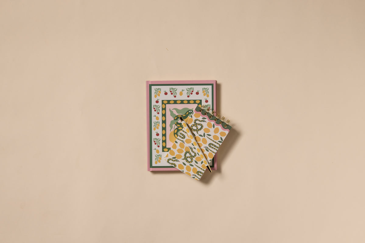 The A Dopo Snake Journal, featuring a Mediterranean-inspired floral cover, rests on a beige surface with two yellow and green patterned cloths partially draped over it.