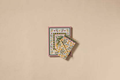The A Dopo Snake Journal, featuring a Mediterranean-inspired floral cover, rests on a beige surface with two yellow and green patterned cloths partially draped over it.