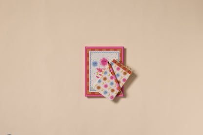 The A Dopo Sun Journal, featuring a vibrant starburst design and lay-flat pages, is paired with a matching notepad and pen, all displayed on a plain beige background.