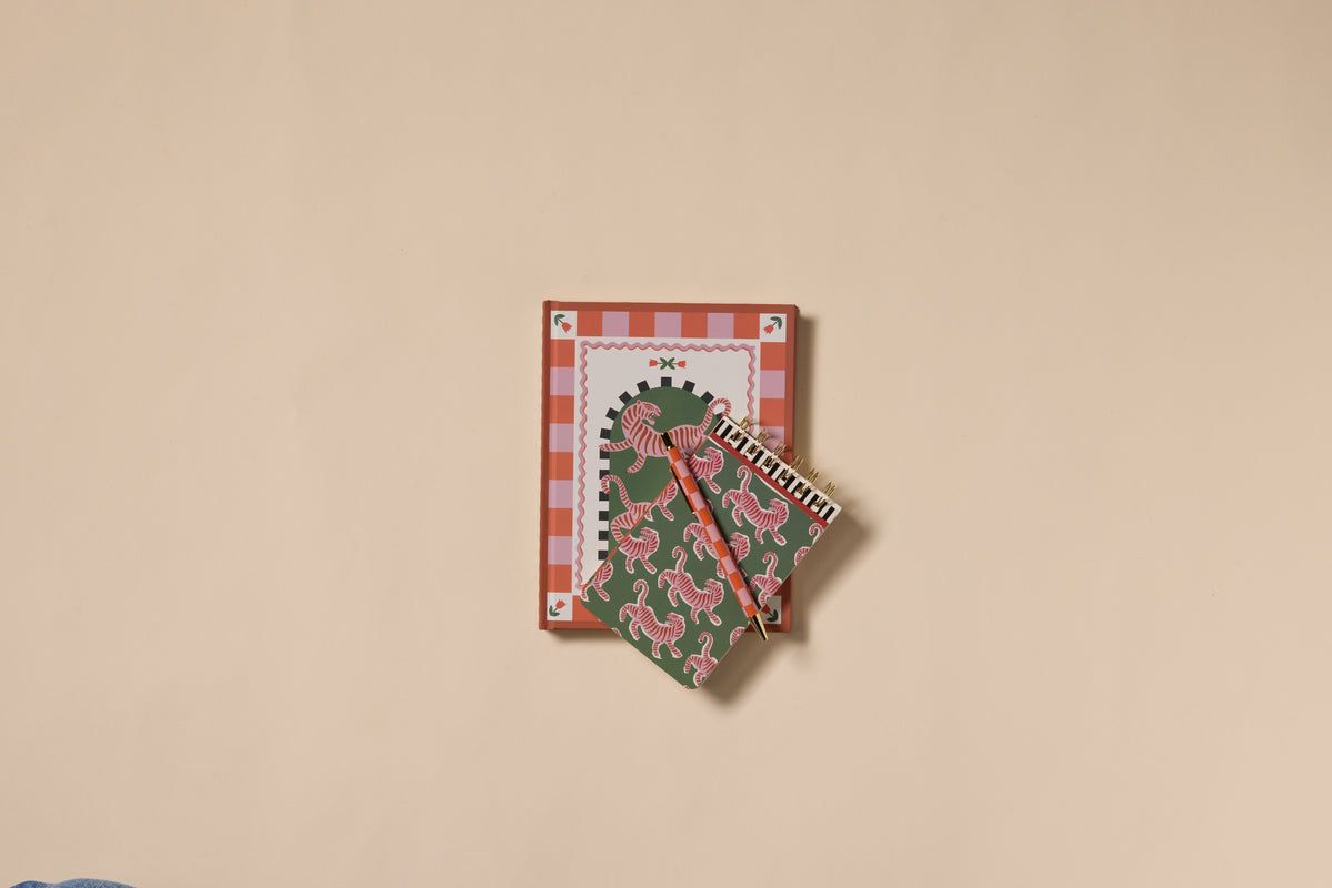 An A Dopo Tiger Journal with lay-flat binding and two spiral notebooks in red and green designs are arranged neatly on a beige background.