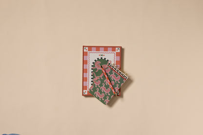 An A Dopo Tiger Journal with lay-flat binding and two spiral notebooks in red and green designs are arranged neatly on a beige background.