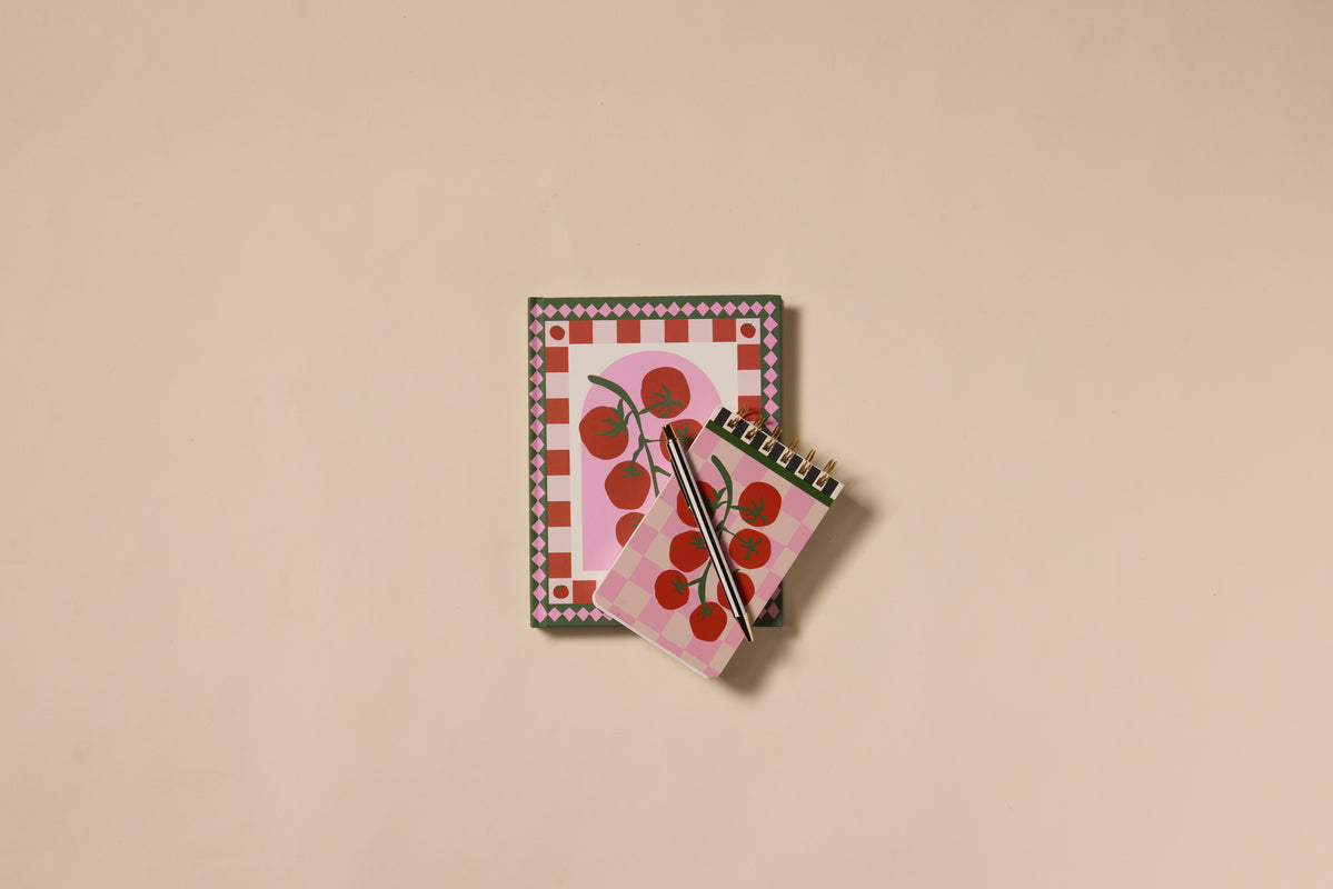 The A Dopo Tomato Journal set includes two lay-flat notebooks with tomato illustrations and checkered patterns, plus a pen—all on a plain beige surface—a perfect choice for your next Italian-inspired journal.