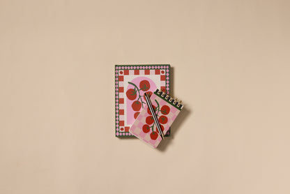 The A Dopo Tomato Twin Wire Notepad set includes two notepads—one large, one small—with hand-drawn red tomato covers on pink and white checkered backgrounds, inspired by Mediterranean travel, plus a black pen on a beige surface.