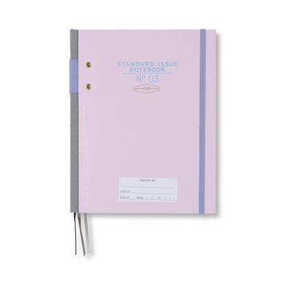 The Standard Issue Planner Notebook No. 03 in Lavender + Periwinkle features a pastel purple cover, a gray spine, elastic closure, two ribbon bookmarks, and a small subject label area at the bottom.
