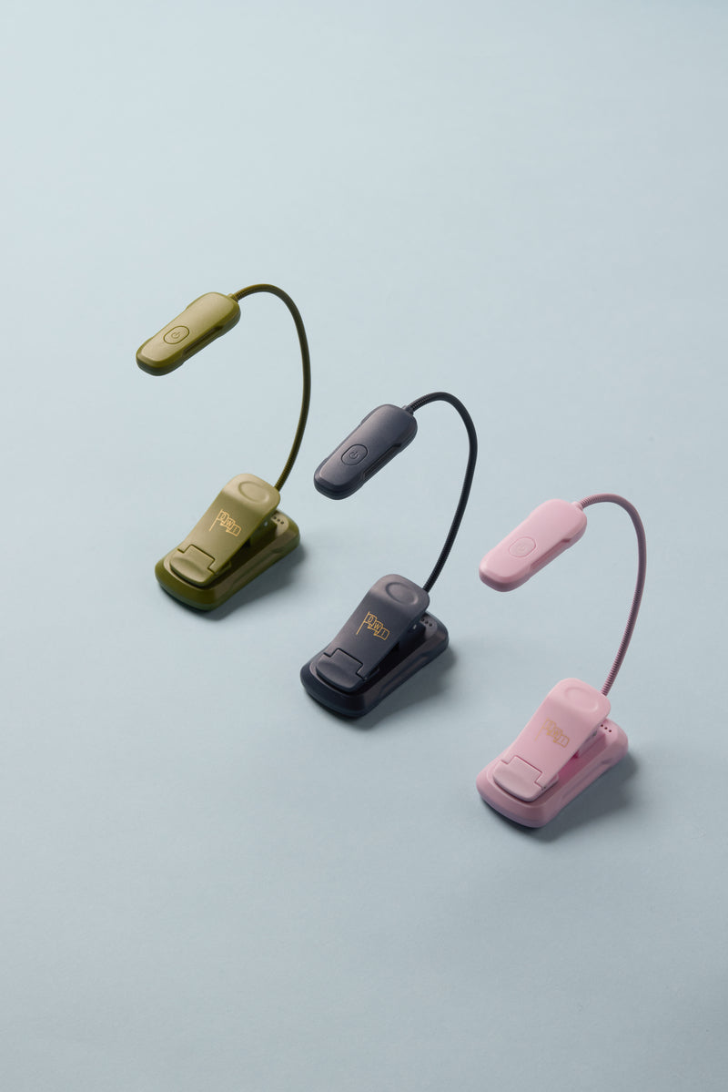 A Book Light - Lilac is arranged on a light blue background. This flexible clip-on LED reading light features a power button and the brand name READ on the base, making it perfect for travel or bedtime reading.