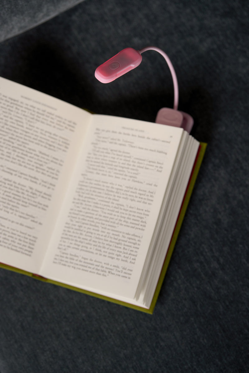 An open book lies on a dark fabric, lit by the Book Light - Lilac, a small flexible LED clipped on top—ideal for late-night reading.