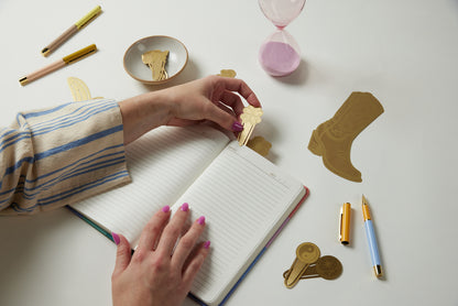 A person with pink nail polish places a gold key-shaped bookmark on an open lined notebook. Pens, book markers, Brass Page Tabs - Lucky Charms, an hourglass, and a small dish rest on the white table.