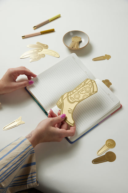 A person places the Brass Bookmark - Cabana Banana, a distinctive book accessory, on an open notebook while other brass bookmarks and pens are scattered nearby on the white table.
