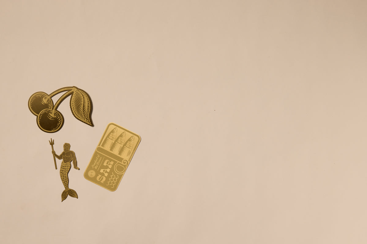 Gold illustrations of cherries, a mermaid with a trident, and sushi on beige cluster in the upper left, creating a refined backdrop—ideal for showcasing the Brass Bookmark - Sardines as an elegant accessory or sturdy placeholder.
