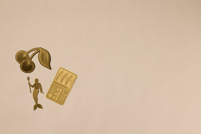 Gold illustrations of cherries, a mermaid with a trident, and sushi on beige cluster in the upper left, creating a refined backdrop—ideal for showcasing the Brass Bookmark - Sardines as an elegant accessory or sturdy placeholder.