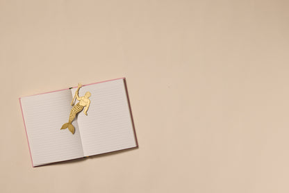 An open pink-covered lined notebook rests on a beige surface with the Brass Bookmark - Poseidon, shaped like a mermaid, clipped to the top left page. The notebook is positioned to the left, leaving empty space on the right.